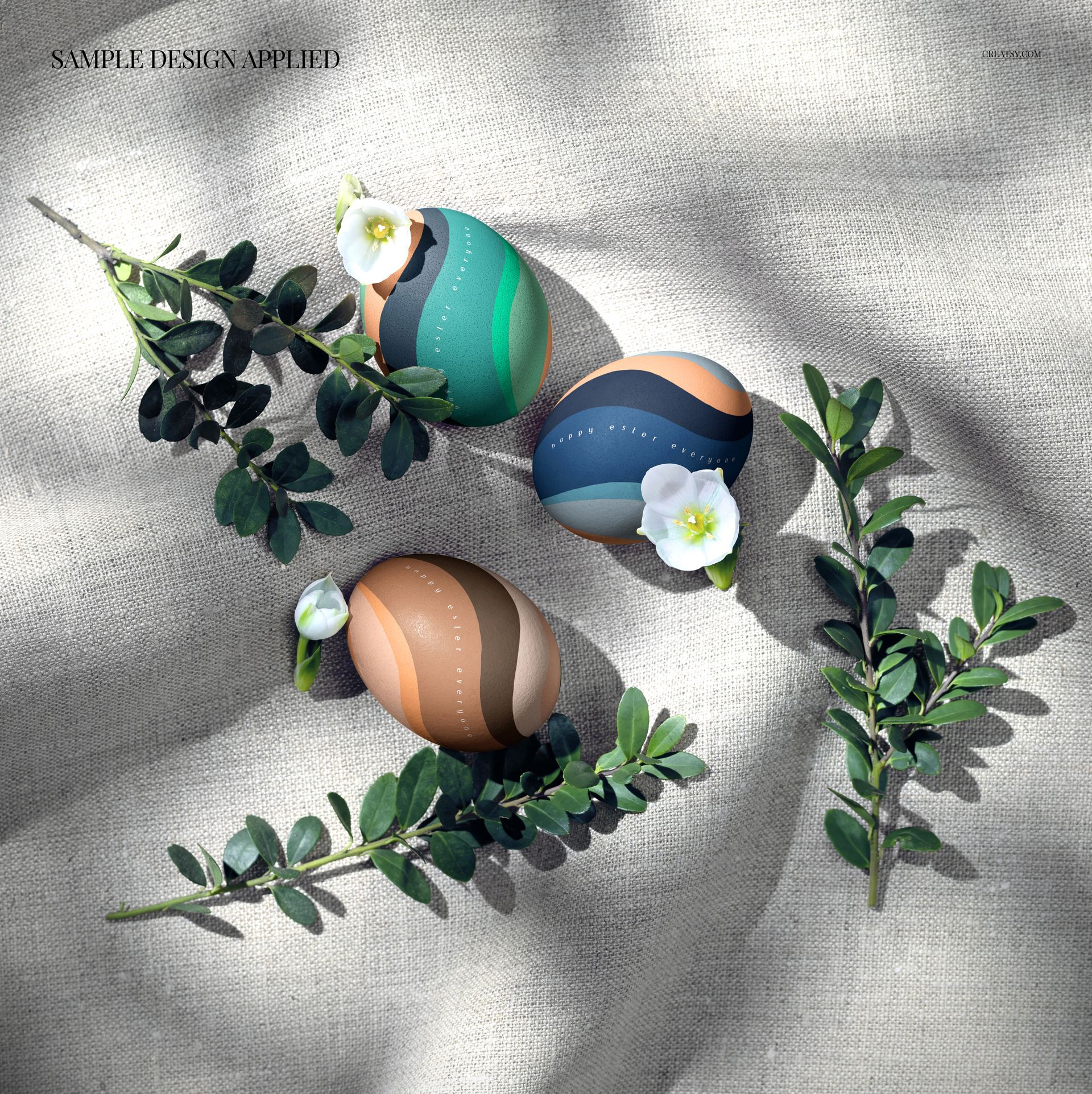 PSD mockup showing three patterned Easter eggs on textured linen, with greenery and delicate white flowers as props.