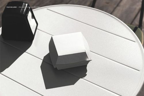Round outdoor table with white napkins holder and closed burger box mockup in bright sunlight, top view.