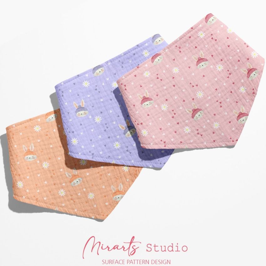 Three baby muslin bandana mockups in pastel orange, purple, and pink, each with a cute rabbit and floral pattern, arranged on a light surface.