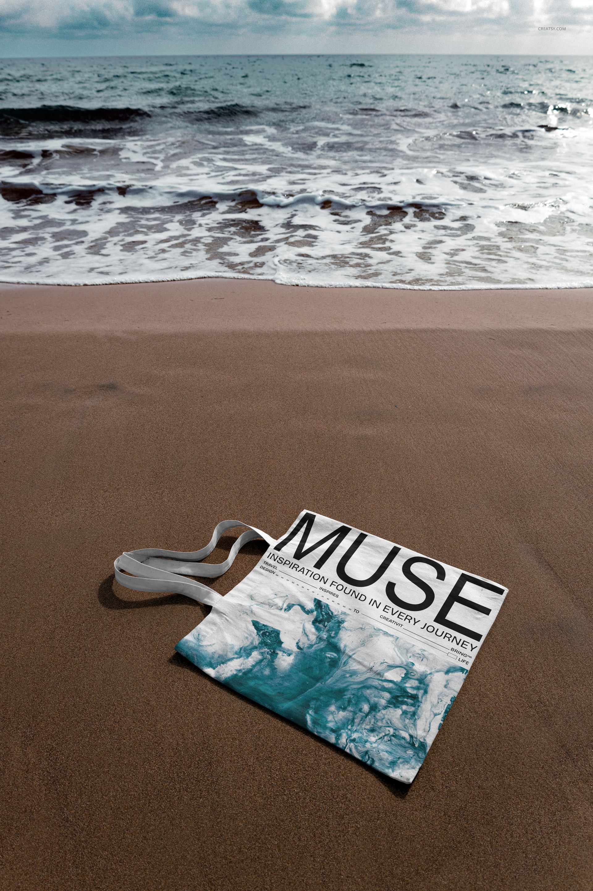 Outdoor PSD mockup featuring a tote bag with white handles and blue design placed on sand by the sea shore.