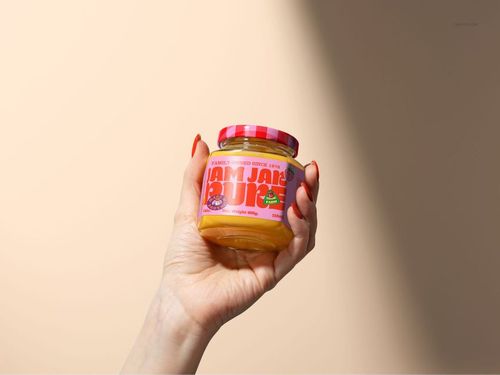 Jam jar mockup shown in a hand, filled with yellow jam, pink patterned lid, and bold text label, studio-lit scene.