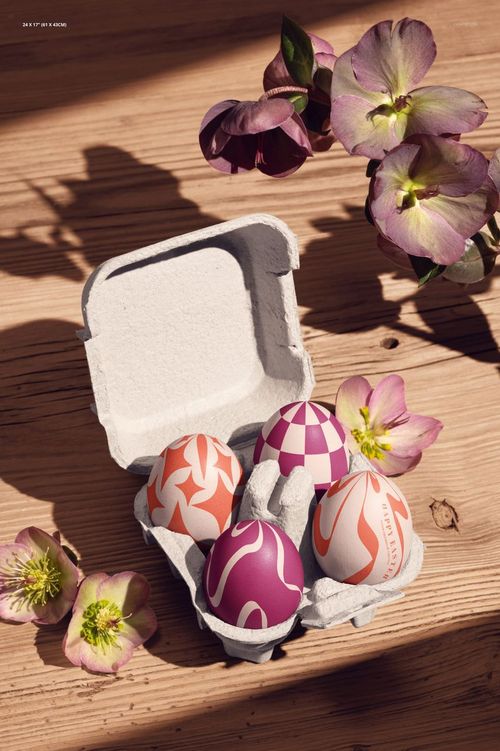 Colorful patterned eggs in a white carton with hellebore flowers arranged around them on a wood table, Easter mockup.