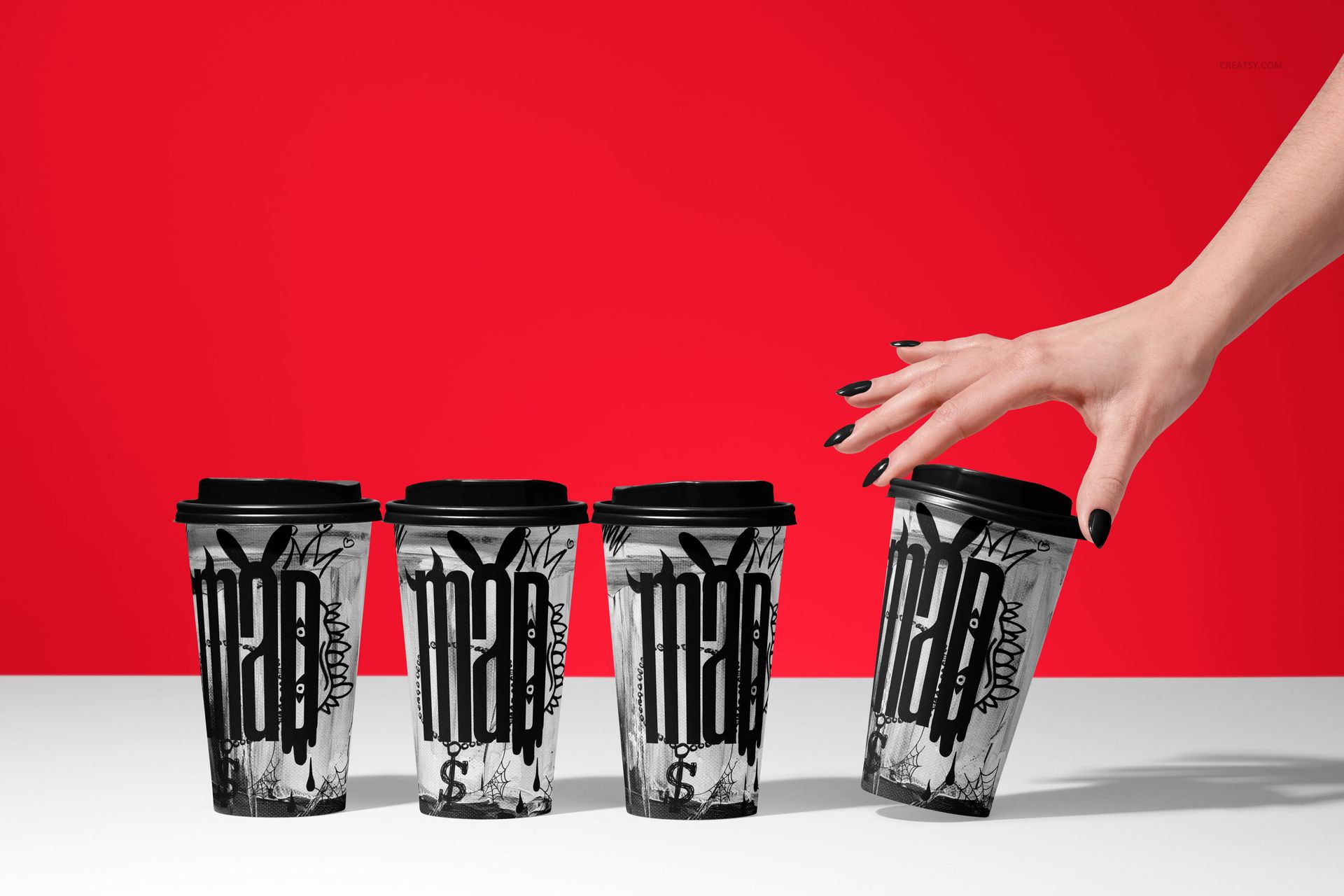 PSD mockup of paper coffee cups featuring black and white graphic prints, with a hand picking up a cup, red backdrop.