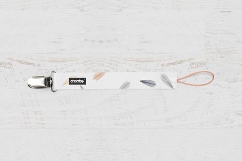 Feather-patterned pacifier clip mockup with editable label, displayed flat on a white wood background.