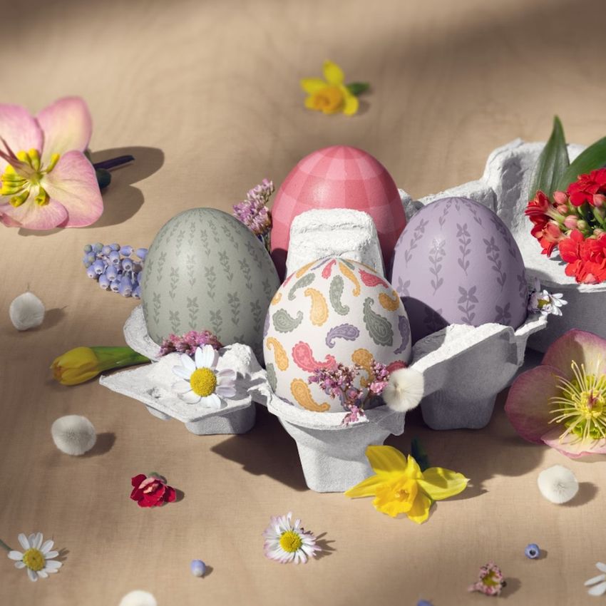 Easter eggs with pastel patterns and paisley designs arranged in a gray carton, surrounded by spring flowers on a light wooden surface.