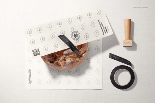 PSD mockup showing a rustic bread loaf wrapped in printed food safe paper, with black tape roll and wooden stamp nearby.
