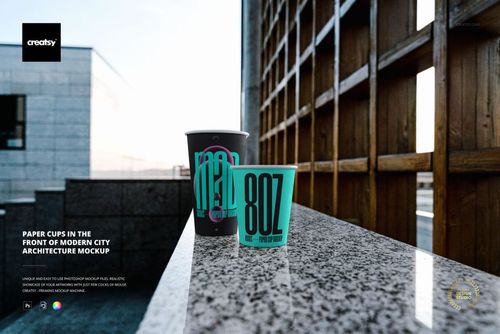 PSD mockup showing paper cups with vibrant lettering, placed outdoors on a stone ledge near contemporary architectural structures.