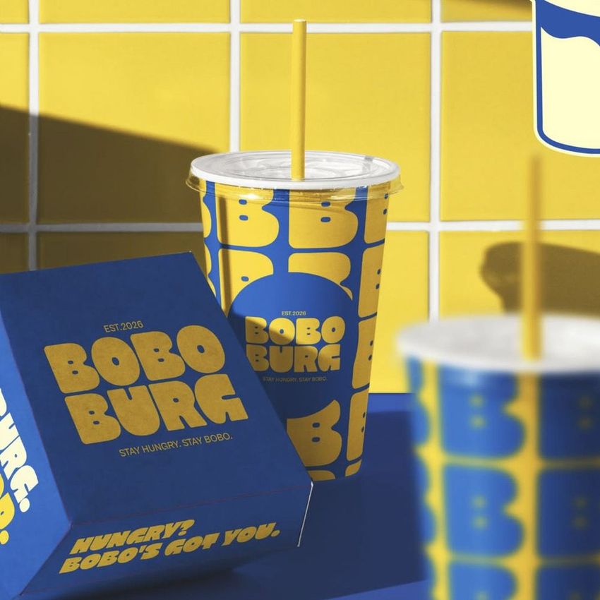 Burger packaging mockup featuring a blue and yellow cup with straw and a matching burger box on a blue surface against a tiled wall.