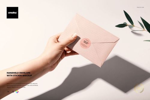Top view of a hand grasping a pink envelope with a sticker closure, stationery mockup scene with plant shadows nearby.