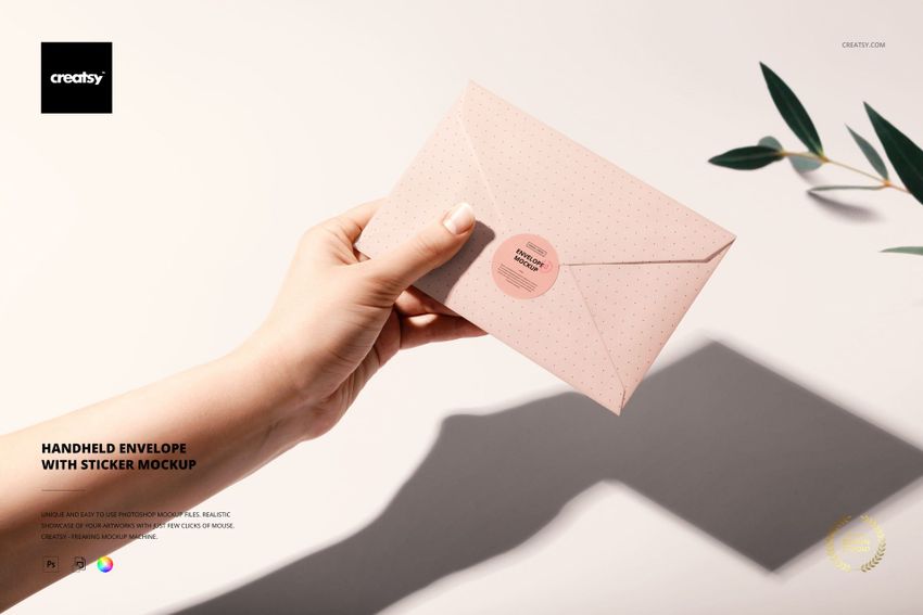 Top view of a hand grasping a pink envelope with a sticker closure, stationery mockup scene with plant shadows nearby.