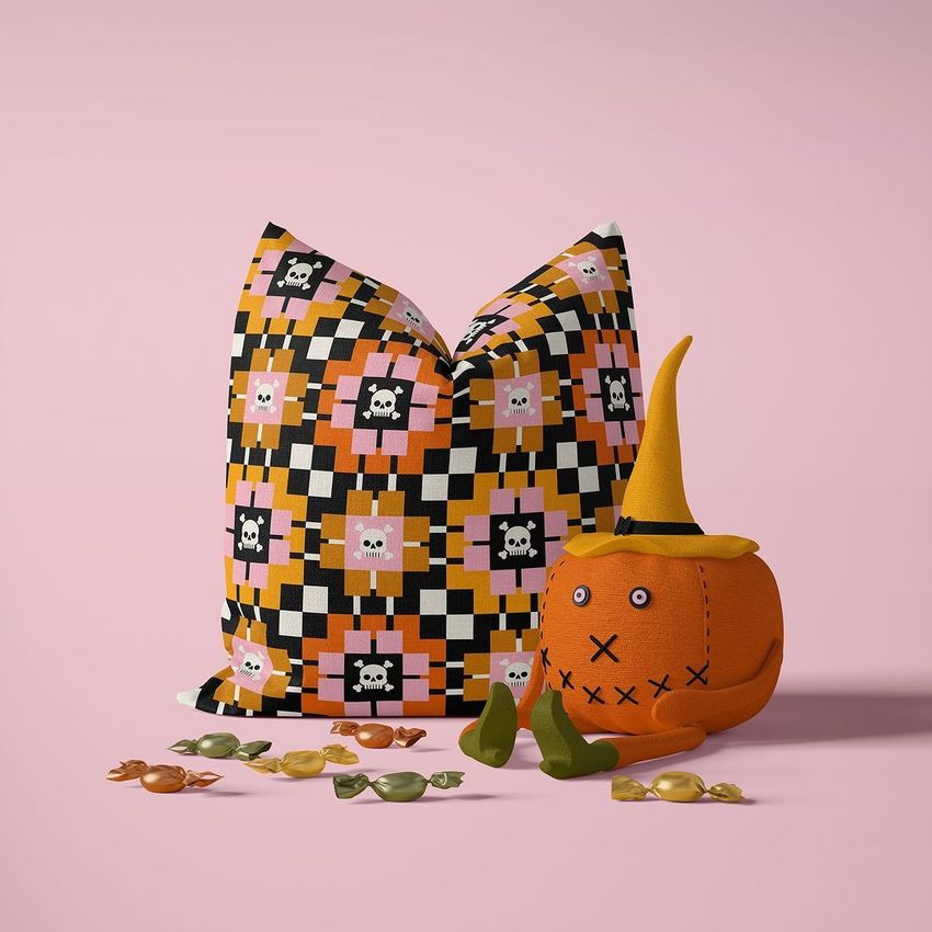 Halloween pillow mockup with a skull and checkerboard pattern in orange, pink, and black, next to a plush toy pumpkin and scattered candies on a light pink background.