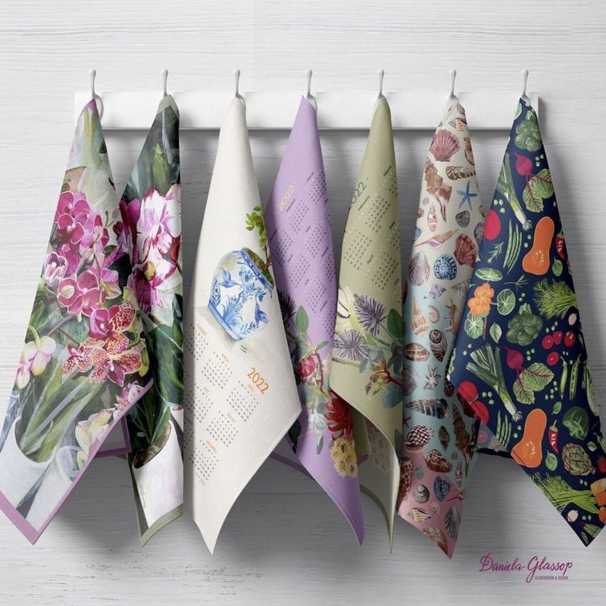Hanging tea towels mockup displaying six towels with botanical and nature-inspired patterns, arranged on white hooks against a light wall.