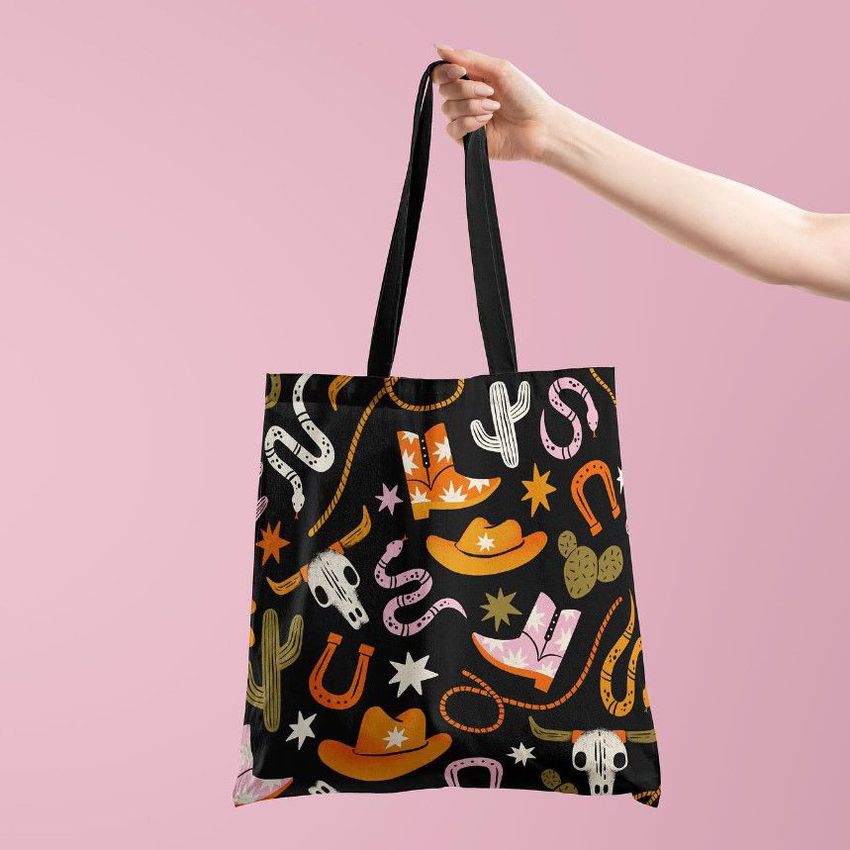 Tote bag mockup featuring a hand holding a black bag with a colorful cowboy-themed pattern on a pink background.