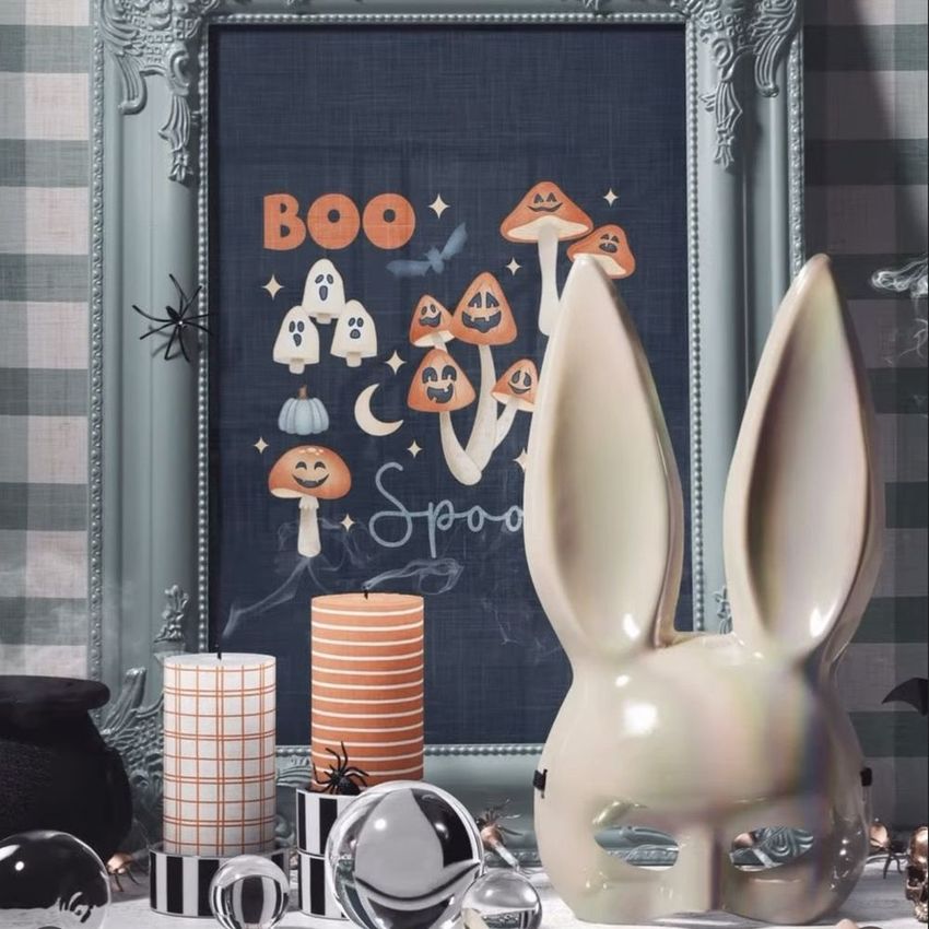 Halloween art print mockup in an ornate pastel frame featuring illustrated mushrooms and ghosts, with candles and ceramic bunny ears in front.
