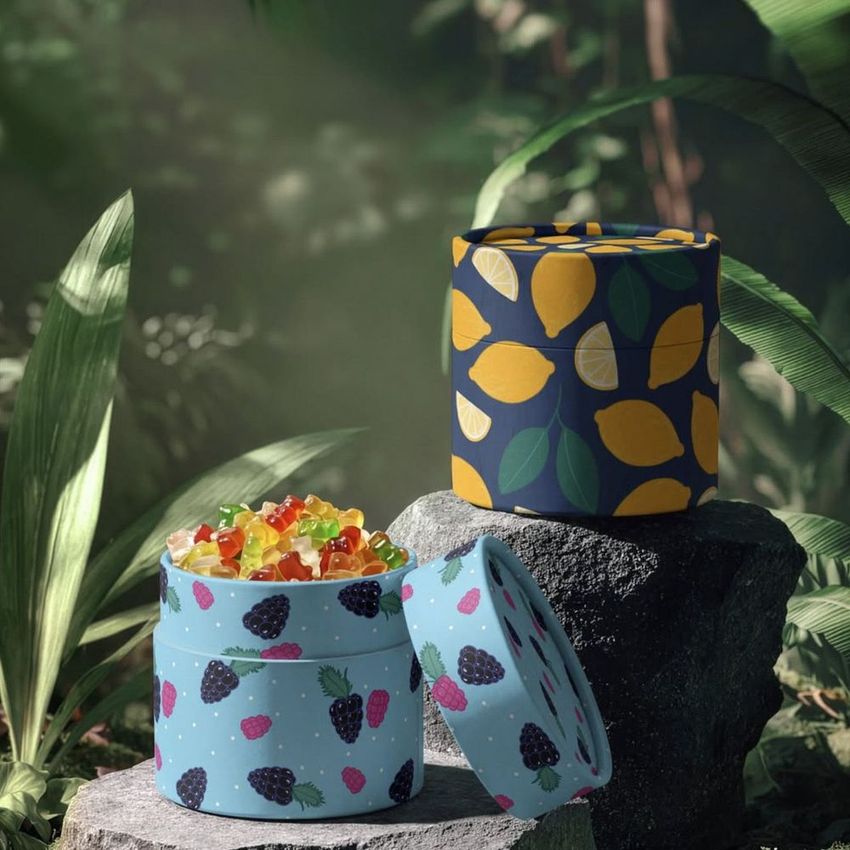 Paper tube packaging mockup featuring fruity patterns, with one open tube filled with gummy candies, set outdoors among plants.