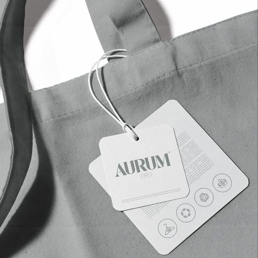 Canvas tote bag mockup in light gray, shown from above with close-up on handle and attached branded tag featuring text and icons.