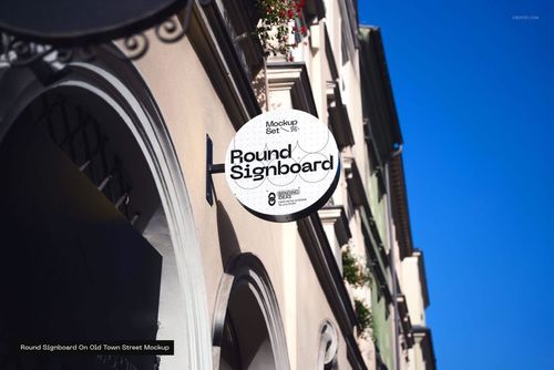 Round signboard mockup mounted on a beige building facade with arched windows in a sunny street scene.