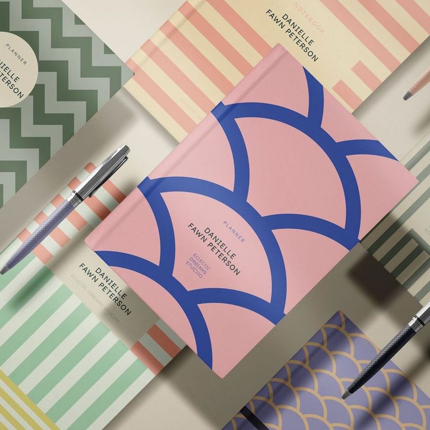 Notebook mockup featuring a pink planner with bold blue geometric patterns, surrounded by pastel stationery and pens on a beige surface.