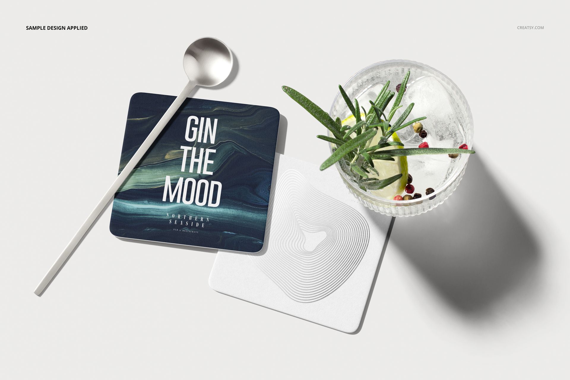 PSD mockup displaying square coasters with modern artwork, accompanied by a clear glass and metallic spoon.