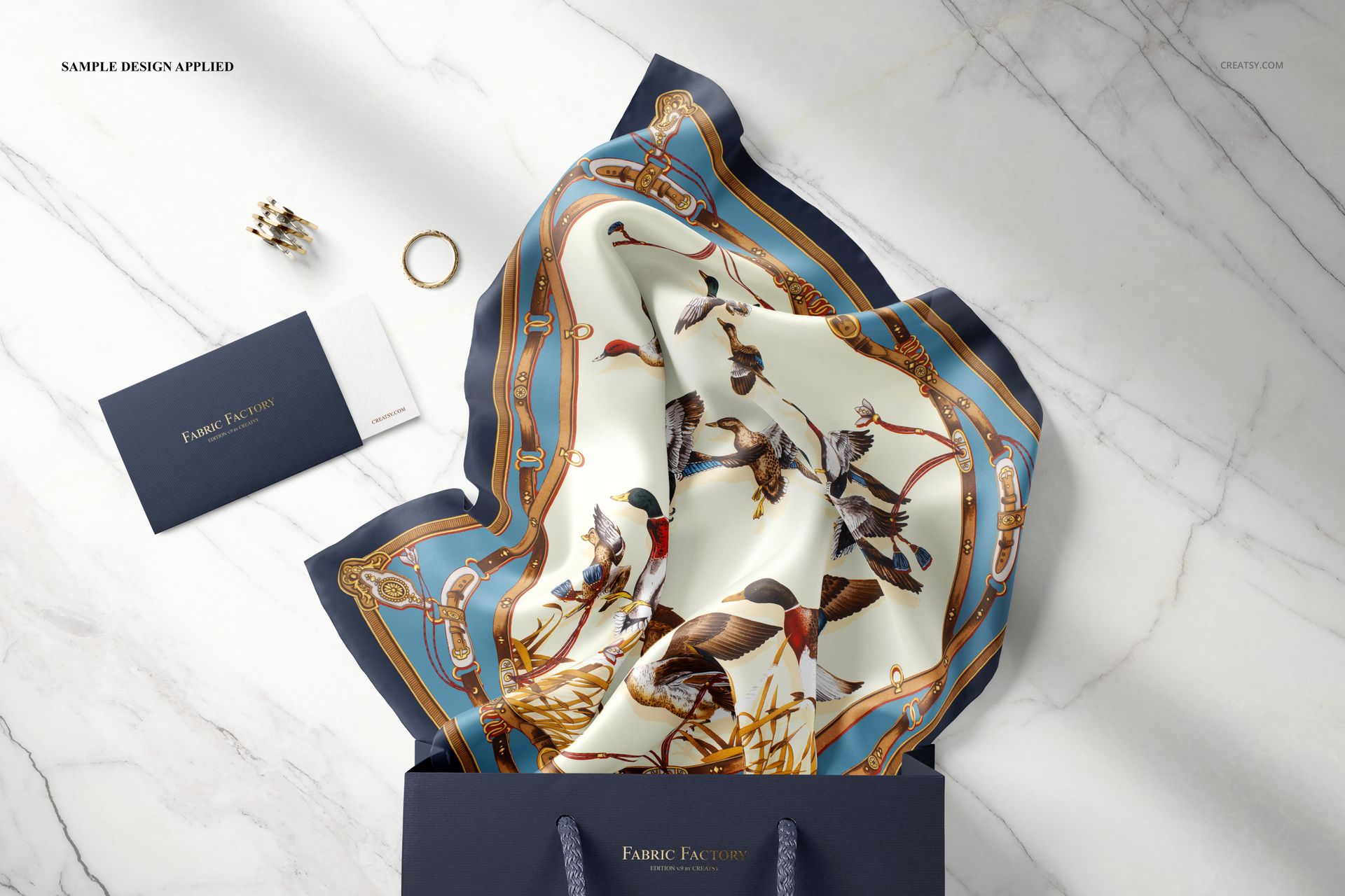 Fashion accessory mockup showing a patterned silk scarf in blue and gold, arranged in a navy bag with jewelry nearby.