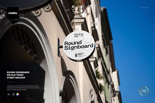 Circular signboard mockup attached to a beige building facade in an old town street, with blue sky above.