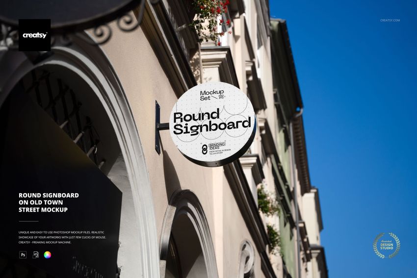 Circular signboard mockup attached to a beige building facade in an old town street, with blue sky above.