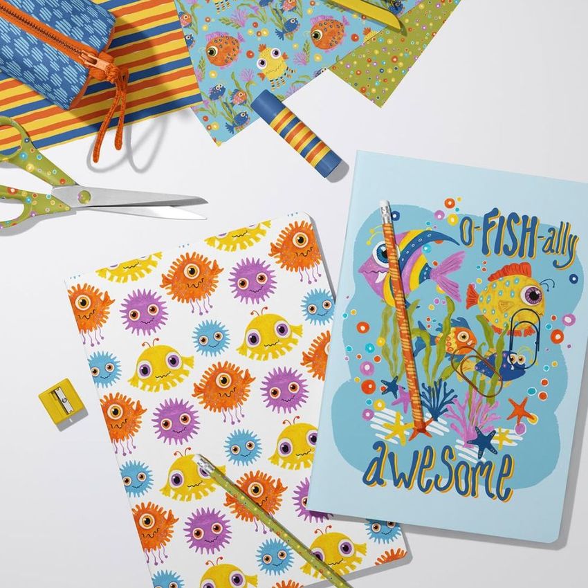 Stationery mockup featuring two A4 notebooks with colorful fish and eye patterns, pencils, scissors, and striped paper on a white surface.