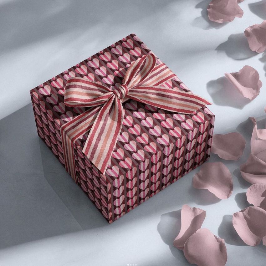 Square gift box mockup with a striped ribbon bow, covered in a pink and red heart pattern, surrounded by scattered rose petals.
