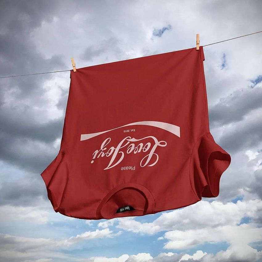 Adult apparel mockup featuring a red t-shirt with visible white text, suspended on a line with clouds in the sky behind.