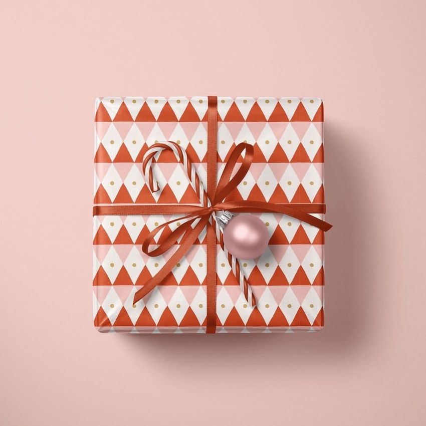 Christmas gift mockup featuring a box wrapped in red and white geometric patterned paper, tied with a ribbon, candy cane, and pink ornament on a light pink background.