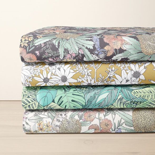 Stack of folded fabric pieces featuring various floral and botanical patterns in muted green, yellow, and black tones on a neutral surface.