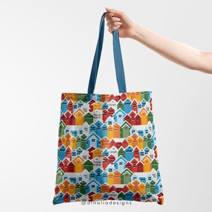 Tote bag mockup featuring a hand holding a bag with a colorful pattern of geometric houses against a plain light background.