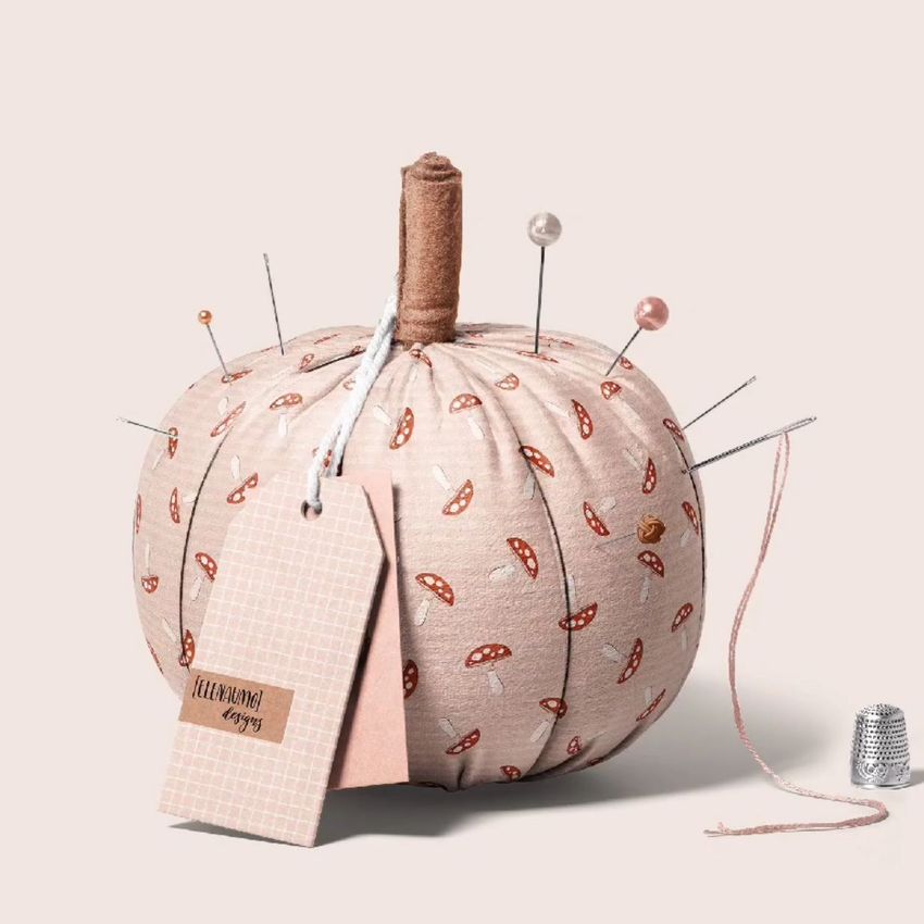 Fabric pumpkin mockup used as a pincushion, decorated with an autumn mushroom pattern, sewing pins, and a hanging tag.
