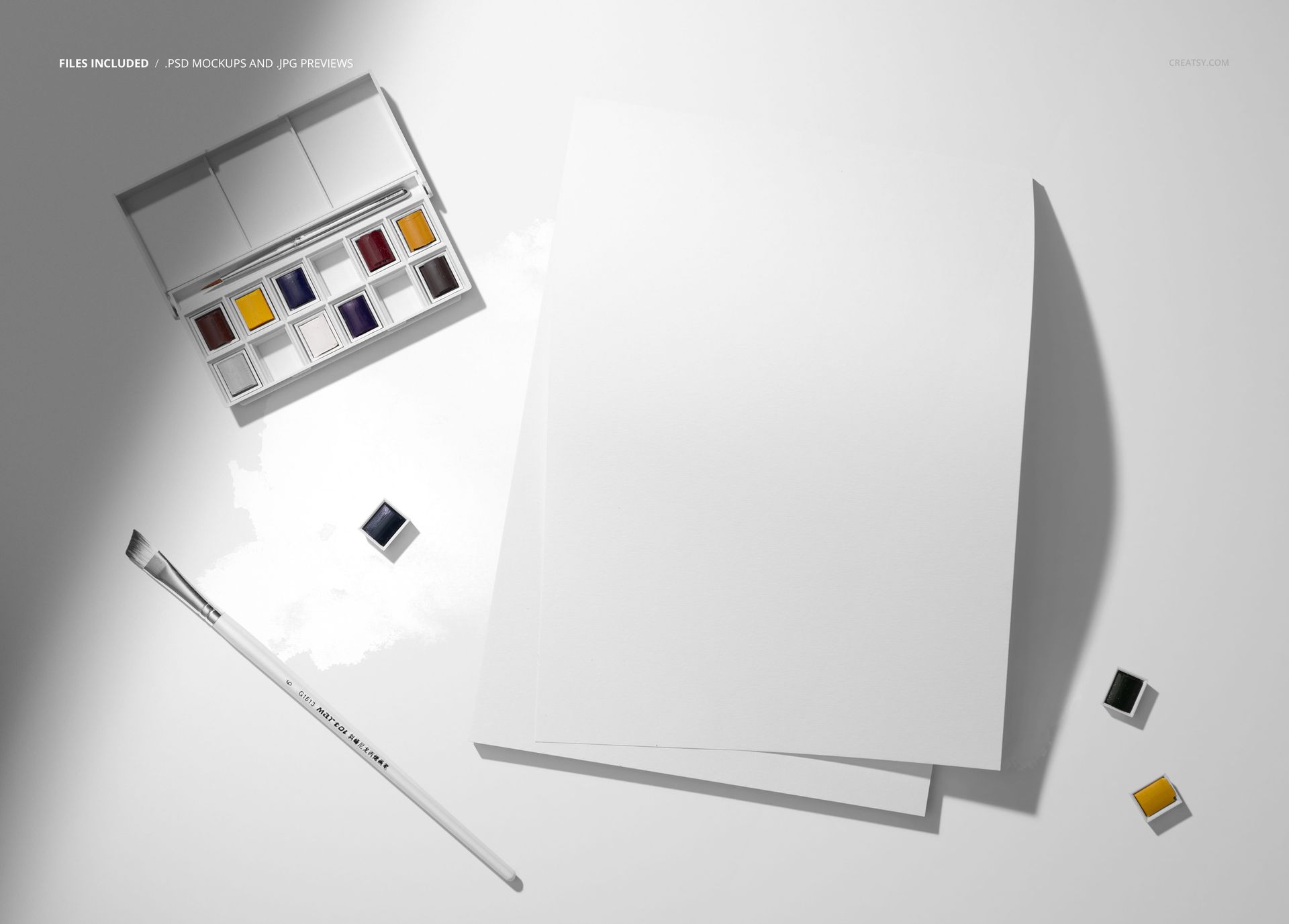 Artistic paper mockup with a watercolor paint palette, paintbrush, and loose paint pans arranged on a flat white background.
