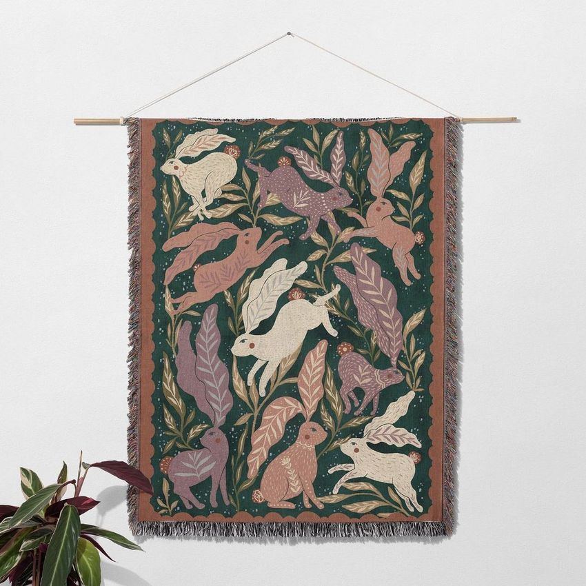 Woven throw blanket mockup featuring a whimsical bunny and leaf pattern in earthy tones, hanging on a wooden rod against a plain wall.
