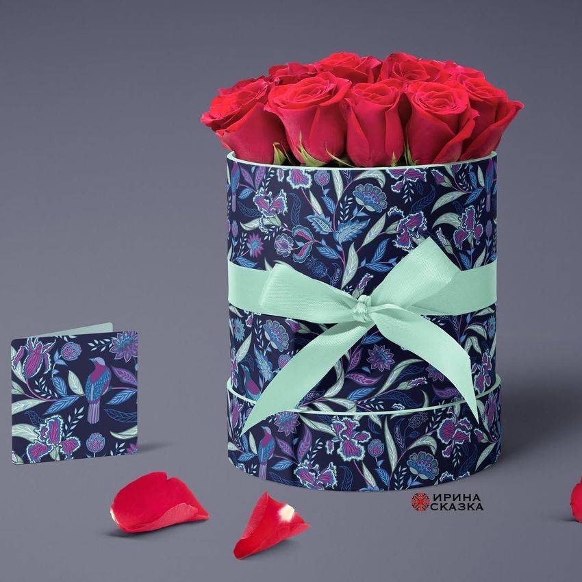 Floral box mockup featuring a cylindrical gift box with a blue and purple botanical pattern, filled with red roses and tied with a light green ribbon.