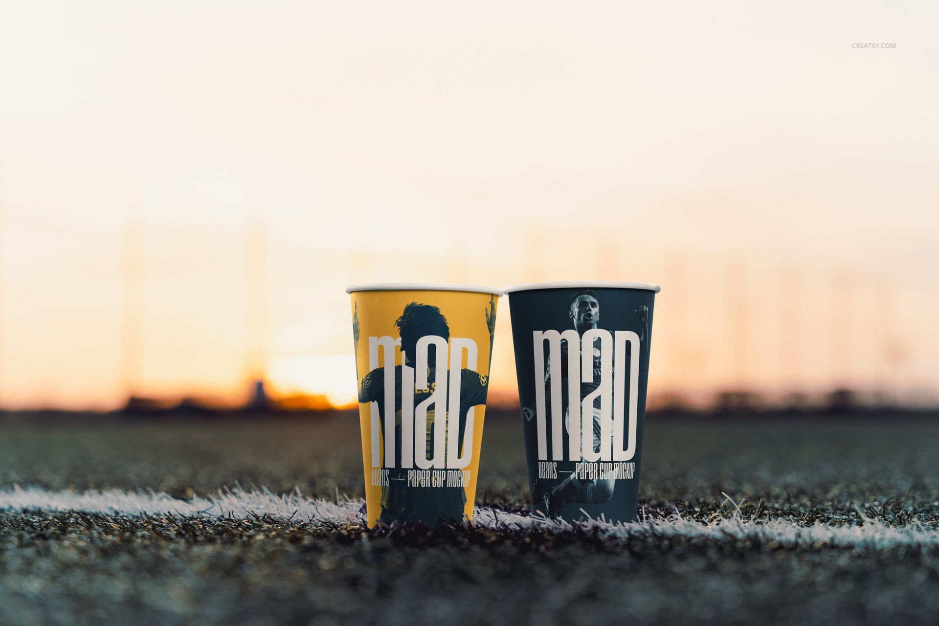 PSD mockup showing a pair of paper cups with artistic prints on a sports field, illuminated by warm evening light.