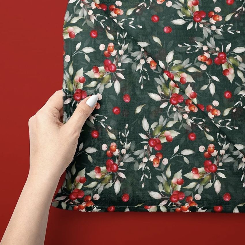 Hand holding a muslin swaddle blanket mockup with a festive Christmas berry and leaf pattern on a dark green background.