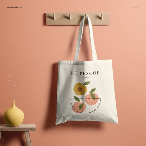 Front view of a white tote bag mockup hanging on a wooden wall rack against a peach-colored wall, featuring a minimalist fruit design.