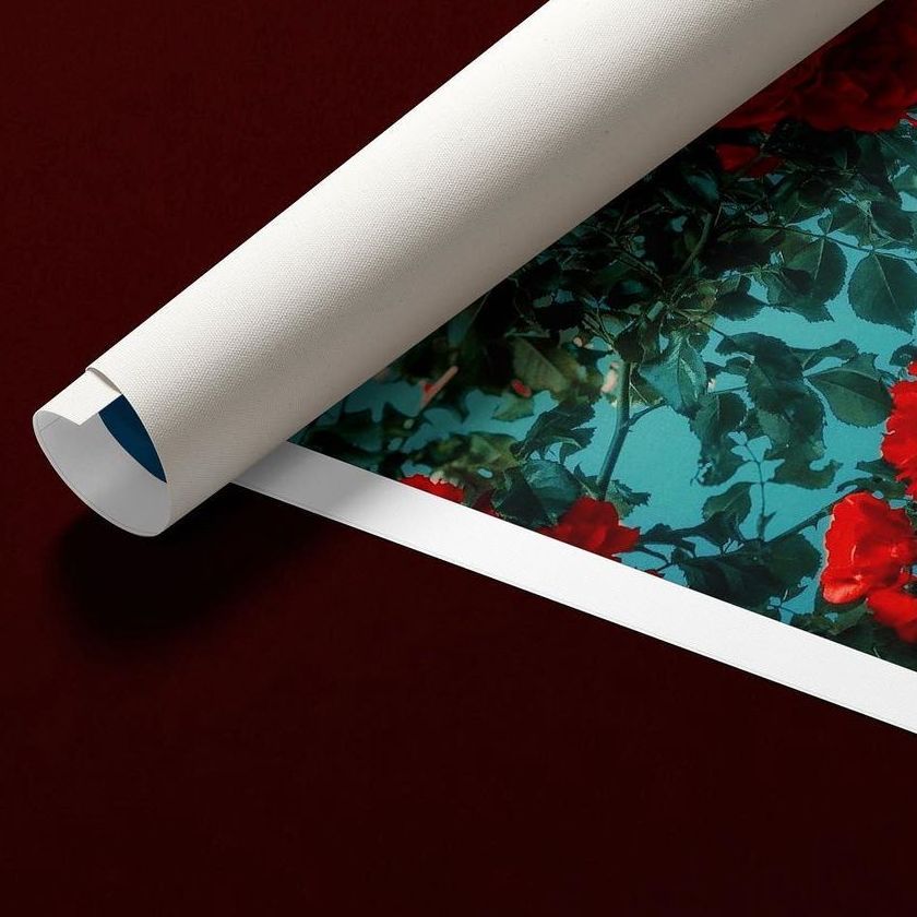 Square rolled canvas print mockup featuring a floral pattern with red flowers and green leaves on a blue background, placed on a dark surface.