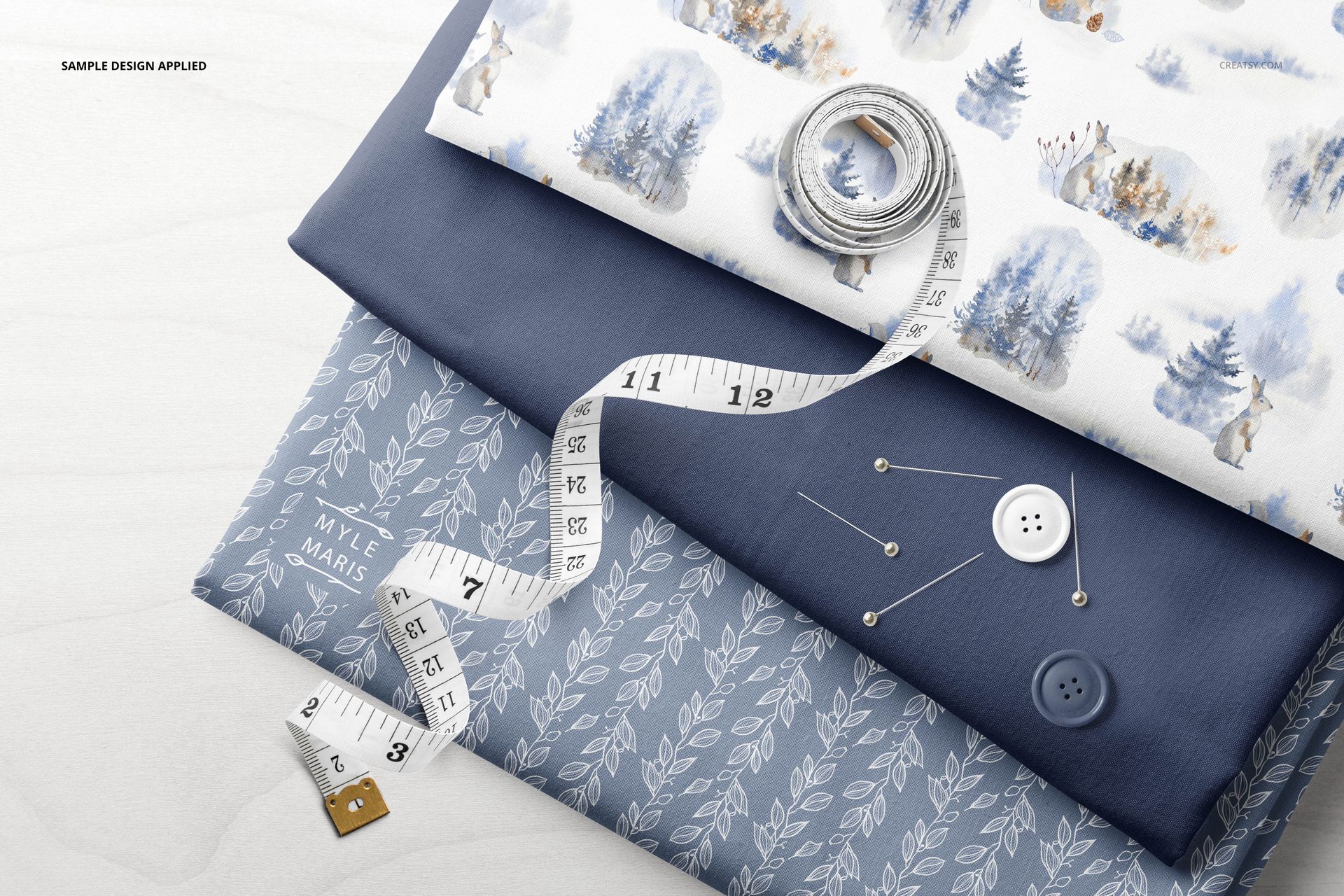 Folded fabrics mockup with blue and white textiles, measuring tape, buttons, and sewing pins arranged on top.