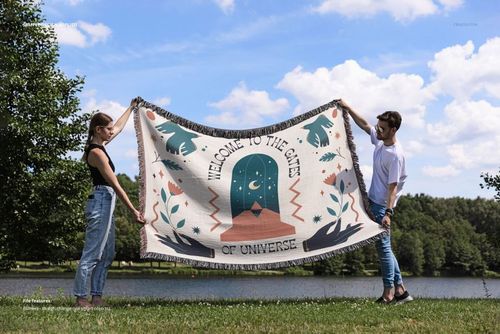 Front view of a fringed woven throw blanket mockup held by two people near a lake, featuring a bold pattern.