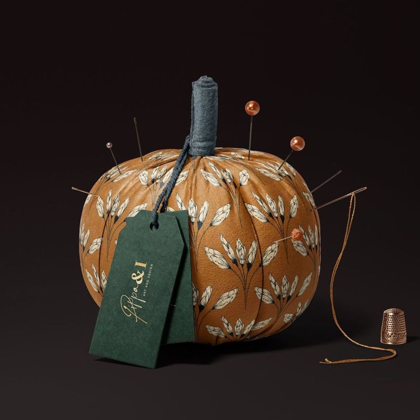 Fabric pumpkin mockup with a brown floral pattern, sewing pins, a green tag, and a thimble on a dark background.