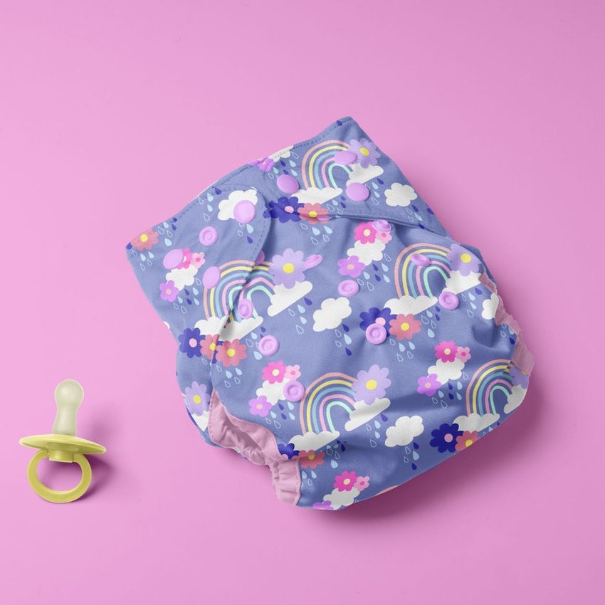 Reusable baby cloth diaper mockup featuring a colorful rainbow and cloud pattern on a purple background, shown with a pacifier on a pink surface.