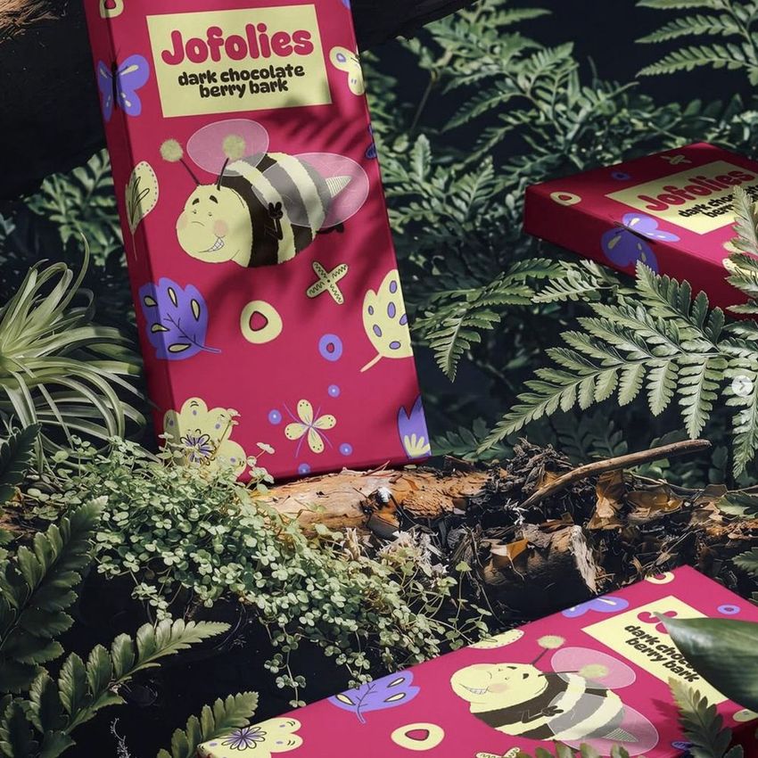 Chocolate box mockup featuring a playful bee illustration on a bright pink package, set among green ferns and forest plants.