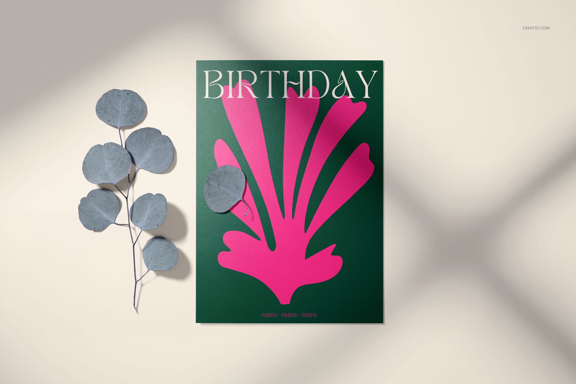 Front view of a 5x7 greeting card mockup with a green cover, pink abstract design, and birthday text, beside dried leaves.