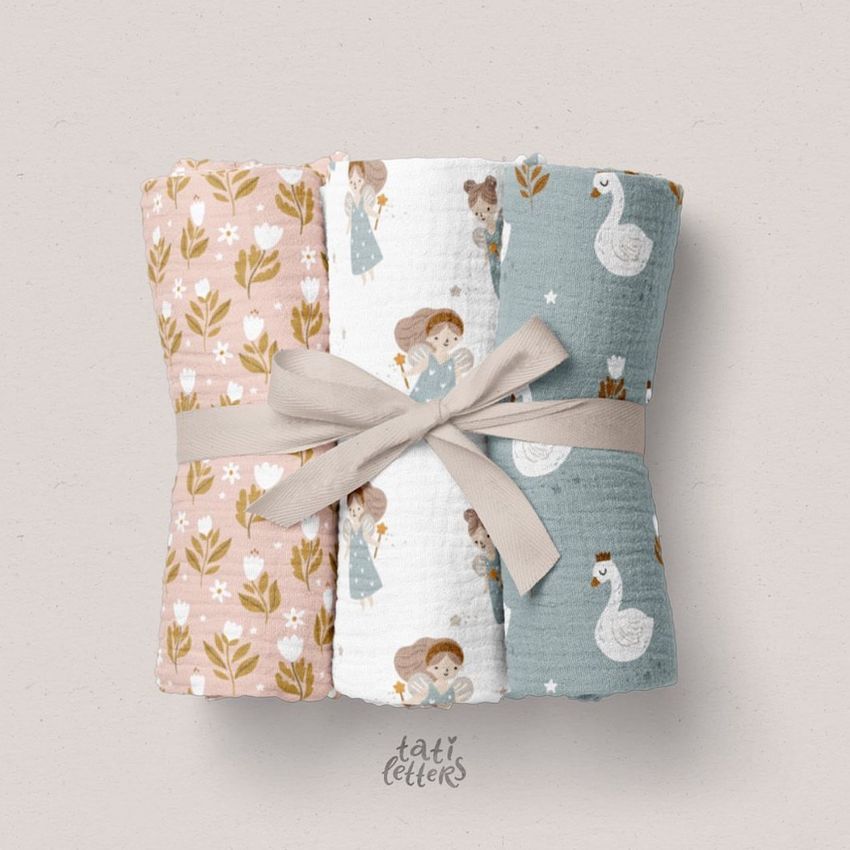 Three crinkle muslin swaddle blankets with pastel floral, fairy, and swan patterns, rolled and tied with a beige ribbon.