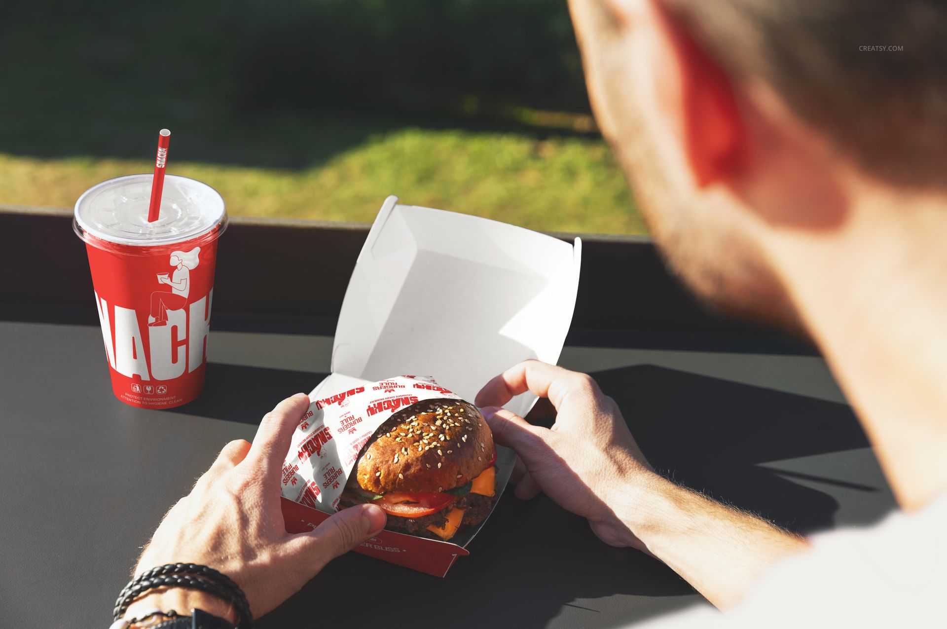 Close-up of a sesame seed burger in a white box, red drink cup with straw, and outdoor background in fast food mockup.