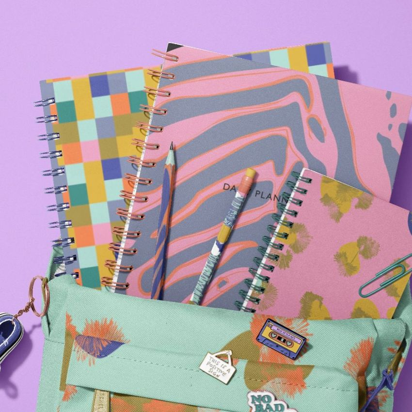 School backpack mockup with spiral notebooks in colorful patterns, pencils, and paper clips on a pastel purple background.