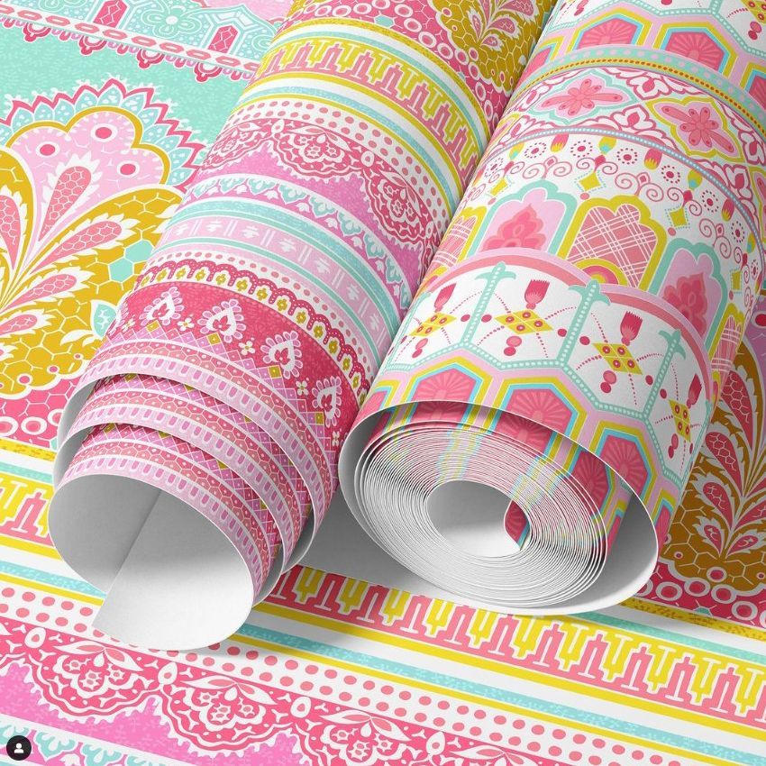 Paper wallpaper mockup showing three rolled sheets with intricate Indian-inspired patterns in pink, yellow, and teal colors.